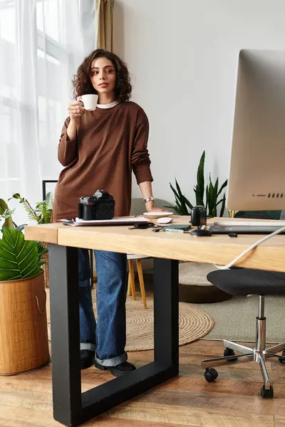 Young woman with curly hair sips coffee at her home office, capturing moments as a photographer. — Stock Photo