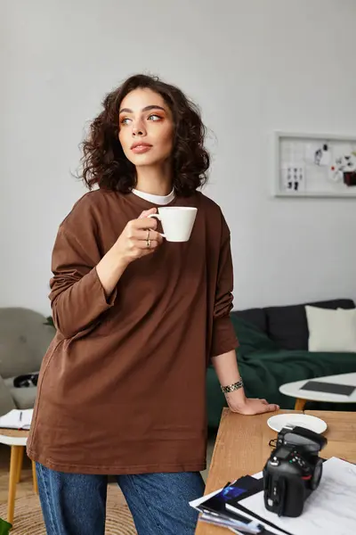 A young woman with curly hair sips coffee, comfortably working from home with her camera nearby. — Stock Photo
