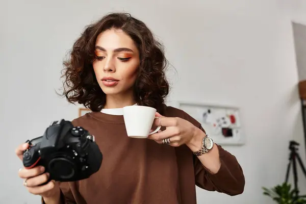 A creative young woman with curly hair sips coffee, focused on her camera while working remotely. — Stock Photo