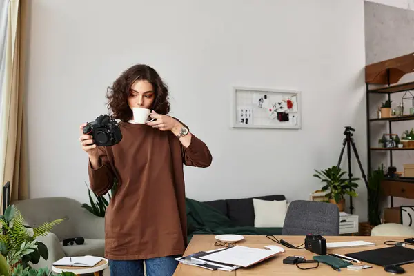 A young woman with curly hair sips coffee while photographing in her cozy home office. — Stock Photo