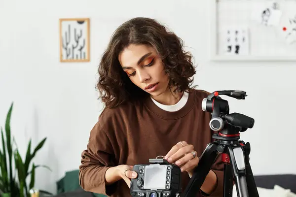 Curly-haired young woman focuses on adjusting her camera while freelancing from her home office. — Stock Photo