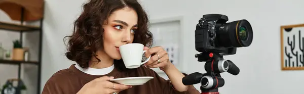 A young woman with curly hair sips coffee and sets up her camera for a creative session at home, banner — Stock Photo