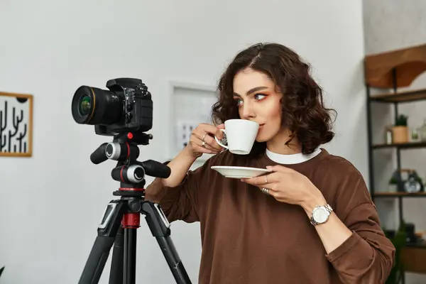 A beautiful young woman is sipping coffee, focused on her camera setup at her cozy home office. — Stock Photo