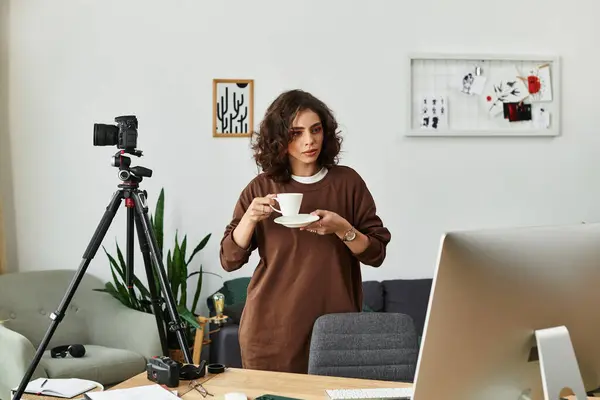 A young woman with curly hair works remotely, sipping coffee as she prepares her camera. — Stock Photo