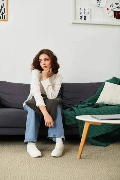 A young woman with curly brown hair sits on a couch, lost in thought in her apartment. — Stock Photo