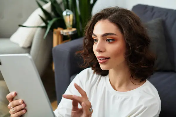Curly brown-haired woman smiles while using a tablet in her modern living space. — Stock Photo