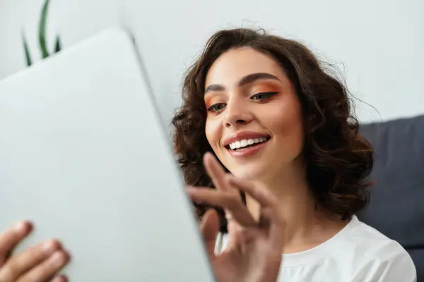 Relaxed in her modern apartment, a young woman with curly hair smiles while using her device. — Stock Photo