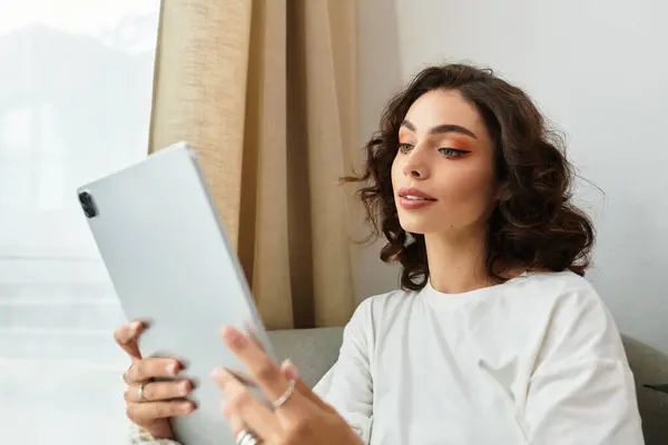 A young woman with curly brown hair relaxes in her modern apartment, using her tablet. — Stock Photo