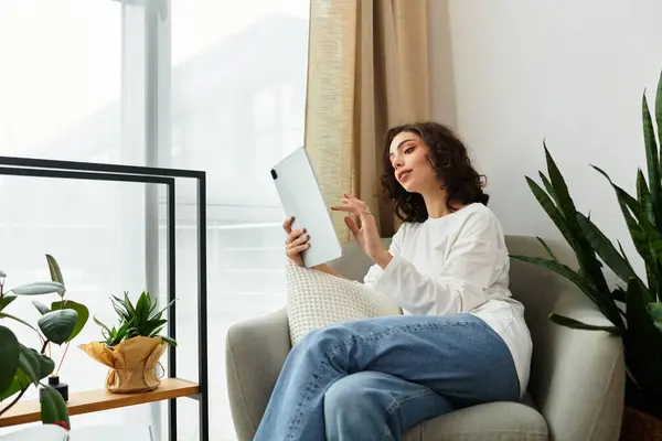 Curly brown-haired woman comfortably rests in her modern apartment while engaging with her tablet. — Stock Photo