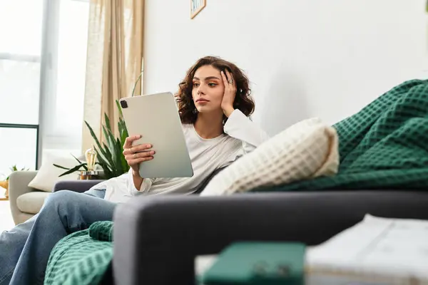 In a modern apartment, a young woman with curly brown hair unwinds while reading on her tablet. — Stock Photo