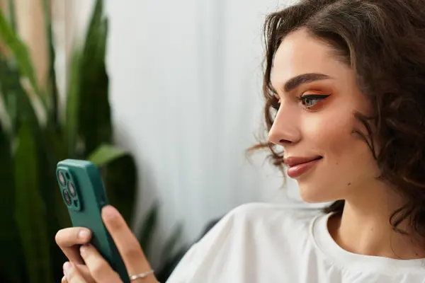 A young woman with curly brown hair relaxes in her stylish apartment while using her smartphone. — Stock Photo