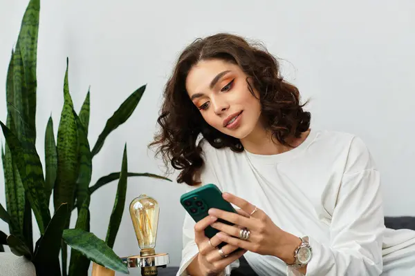 Young woman with curly hair relaxes on a cozy couch, using her smartphone among plants. — Stock Photo