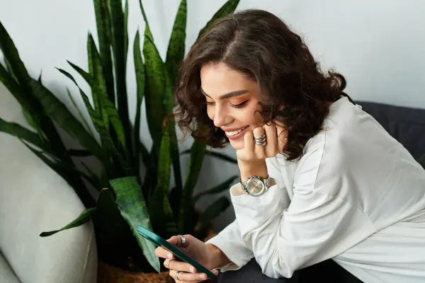 Curly brown-haired woman relaxes in her stylish apartment, focused on her phone and smiling. — Stock Photo