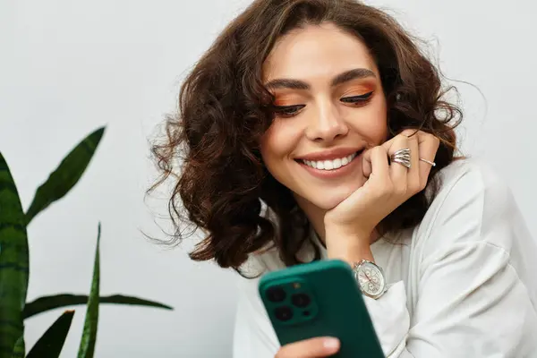 A young woman with curly brown hair smiles while relaxing in her modern apartment, using her phone. — Stock Photo