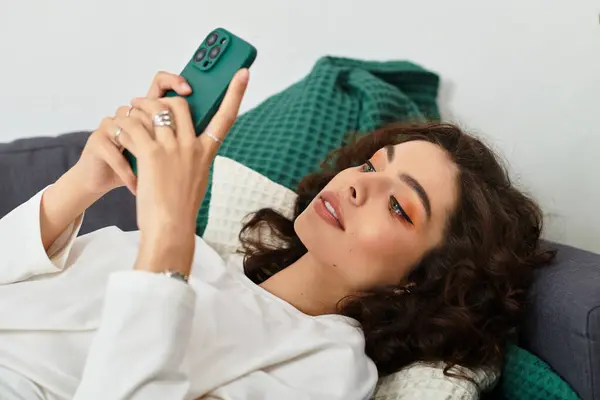 A beautiful young woman with curly brown hair rests on a couch, engaged with her smartphone. — Stock Photo