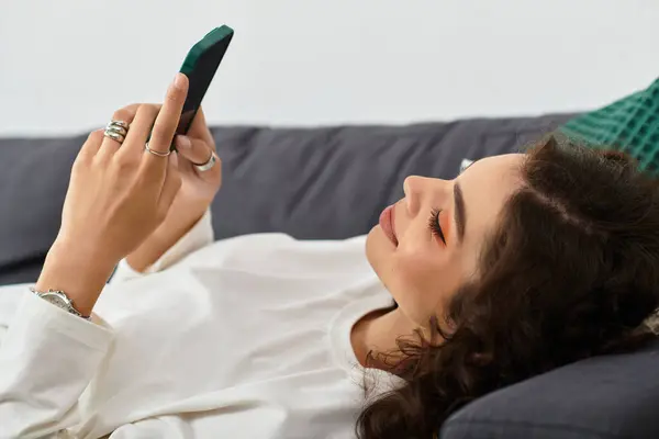 A beautiful young woman with curly brown hair relaxes calmly in her trendy apartment while texting. — Stock Photo