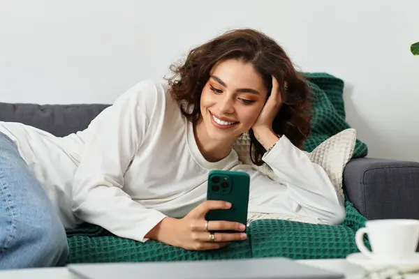 A beautiful young woman with curly brown hair rests on a cozy couch, smiling at her phone. — Stock Photo