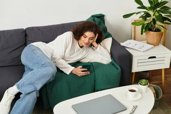 Young woman with curly hair relaxes on a couch, engaging with her smartphone for remote work. — Stock Photo