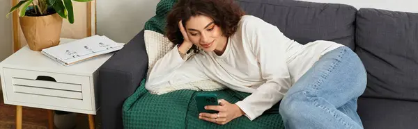 Curly-haired woman rests on a couch in her modern apartment, absorbed in her phone, banner — Stock Photo