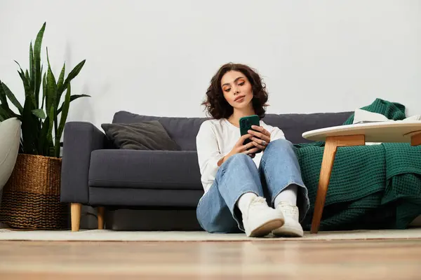 A young woman sits comfortably on the floor of her modern apartment, focused on her smartphone. — Stock Photo