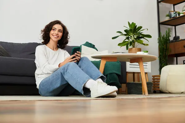 A young woman with curly hair sits relaxed on the floor of her stylish apartment, on her phone. — Stock Photo