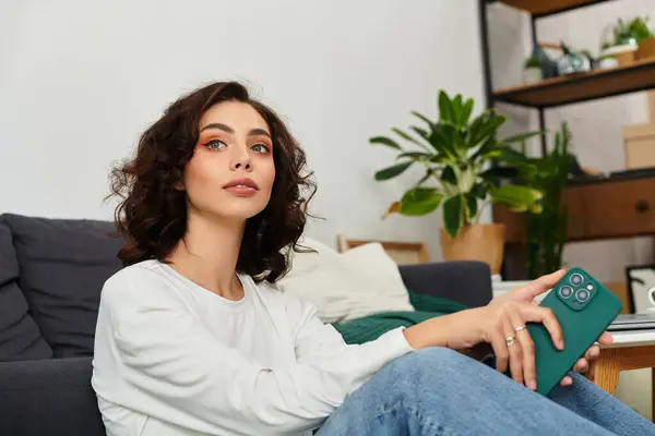 Young woman with curly brown hair relaxes in her stylish apartment, holding a phone. — Stock Photo