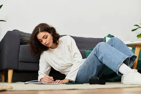 Young woman with curly brown hair lies on the floor, writing in her notebook in a stylish apartment. — Stock Photo