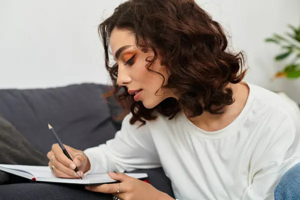 Curly-haired woman is focused on writing in her journal while relaxing at home in a stylish setting. — Stock Photo