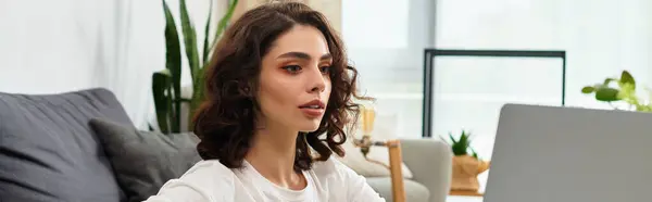 A young woman with curly hair focuses intently on her laptop in a bright and inviting room, banner — Stock Photo