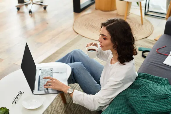 A young woman with curly hair focuses on her laptop while having coffee in a comfortable setting. — Stock Photo