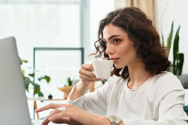 A young woman with curly hair sips coffee, focused on her remote work at home. — Stock Photo