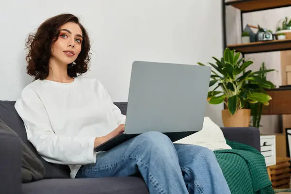 Young woman with curly hair sits comfortably on a couch, working from home on her laptop. — Stock Photo