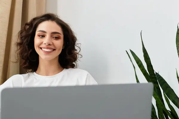 Young woman with curly hair smiles while working on her laptop from a cozy home environment. — Stock Photo