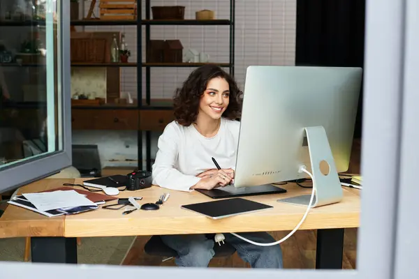 A young woman with curly hair joyfully edits and retouches images using her graphic tablet at home. — Stock Photo