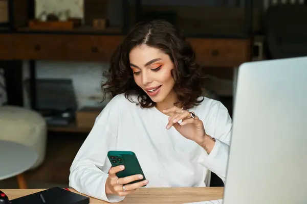 A beautiful young woman with curly hair is focused on her smartphone while working from home. — Stock Photo