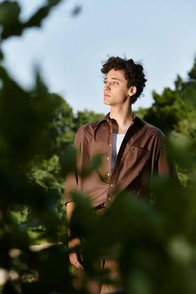 Curly-haired young man relaxes outdoors in a fashionable summer outfit surrounded by greenery. — Stock Photo