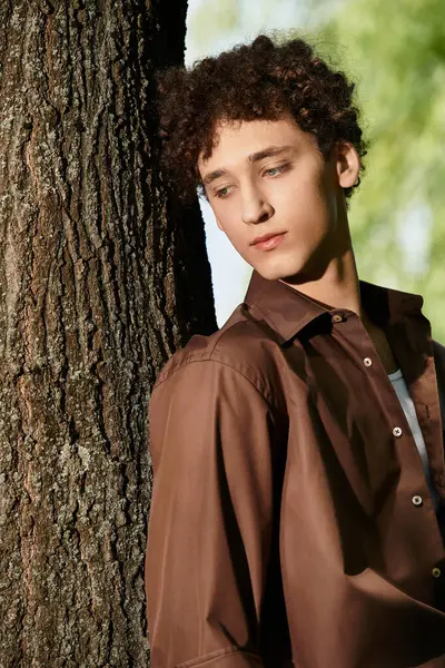 A stylish young man with curly hair relaxes against a tree in a lush green park. — Stock Photo