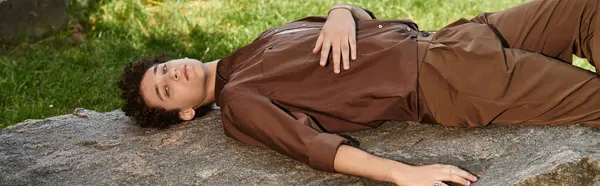 Young man with curly hair enjoys a sunny day while lounging on a rock in the park, banner — Stock Photo