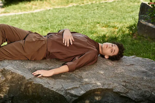 A young man relaxes on a large rock, dressed in a trendy summer outfit amid lush greenery. — Stock Photo