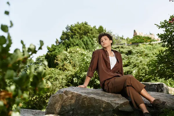 A young man with curly hair relaxes on a rock in a vibrant park, embracing summer vibes and nature. — Stock Photo