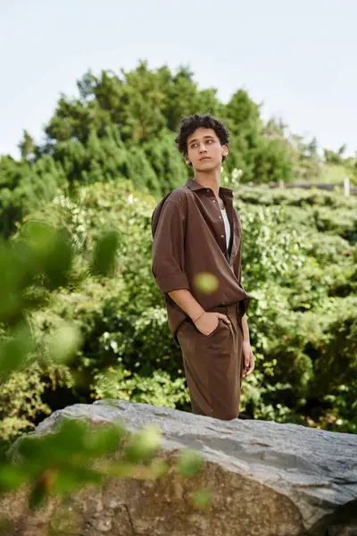 Curly-haired young man in a fashionable outfit stands on a rock, soaking up the sun in a park. — Stock Photo
