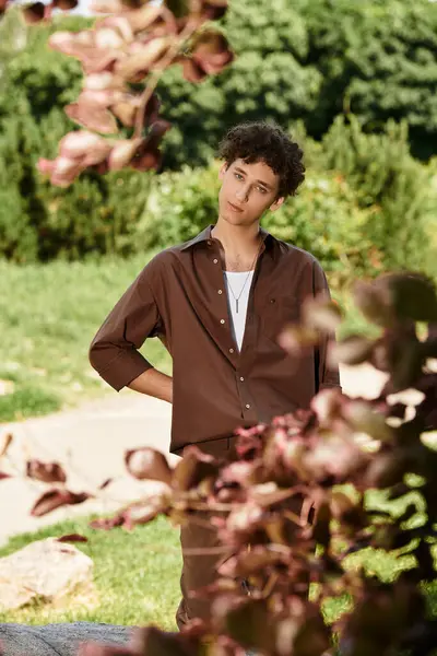 Curly-haired young man relaxes in a lush park, dressed in a trendy summer outfit, embodying leisure. — Stock Photo