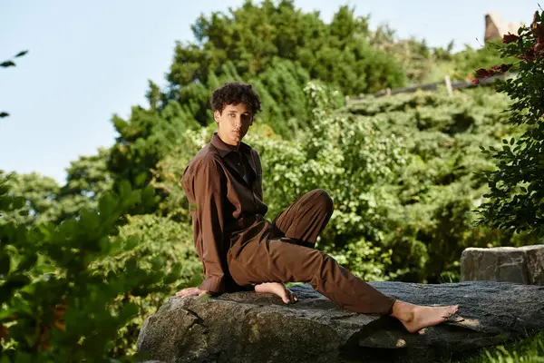 A young man with curly hair relaxes barefoot on a rock in a lush park, soaking up the sun. — Stock Photo