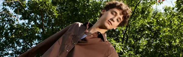 A young man with curly hair relaxes in a green park, embracing the joy of summer sunshine, banner — Stock Photo
