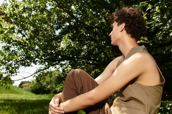 Curly-haired young man relaxes outdoors in a stylish outfit, soaking up the warm sunshine. — Stock Photo