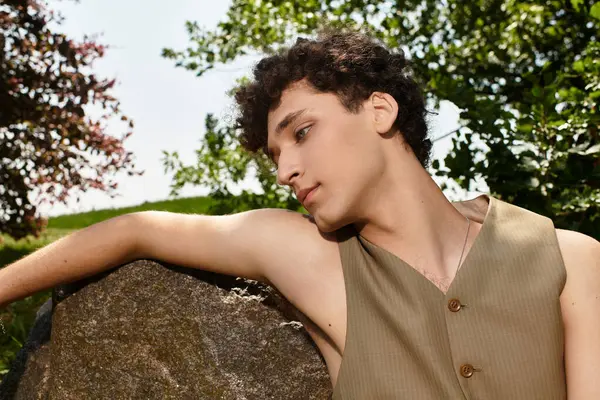 Curly-haired young man relaxes in a lush park, showcasing his stylish summer look amidst nature. — Stock Photo