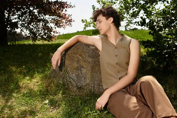 A young man with curly hair relaxes on a stone in a lush park, showcasing summer fashion. — Stock Photo