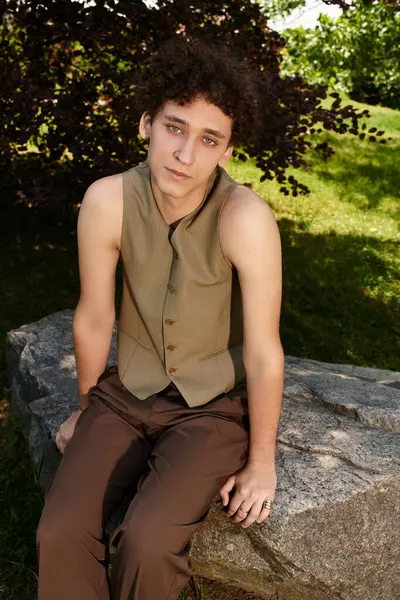 A young man with curly hair lounges in the park, wearing a stylish summer outfit. — Stock Photo