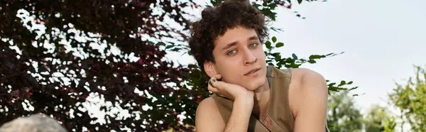A young man with curly hair relaxes under the sun in a lush green park, embracing summer vibes, banner — Stock Photo