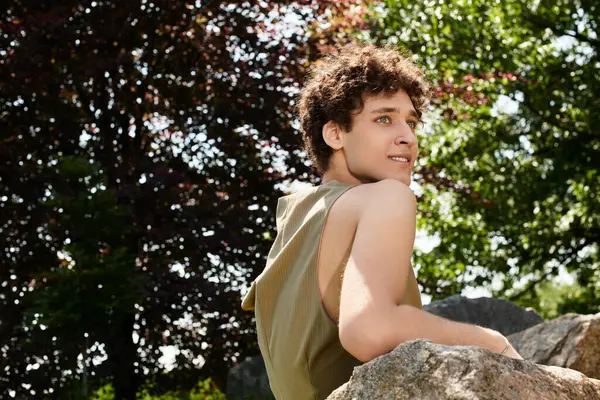 Curly-haired young man relaxes on a rock in a vibrant park surrounded by greenery under blue skies. — Stock Photo